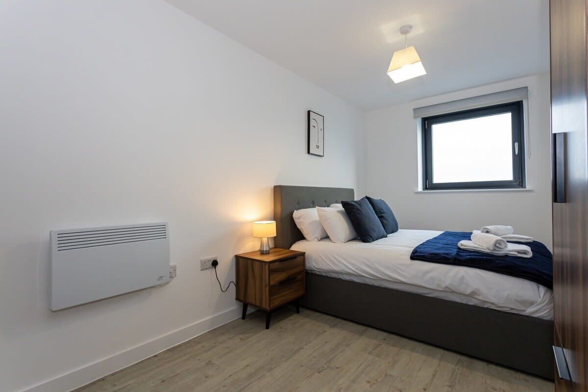 Ein Schlafzimmer mit Bett, Nachttisch, Lampe, Fenster und Wandheizung.