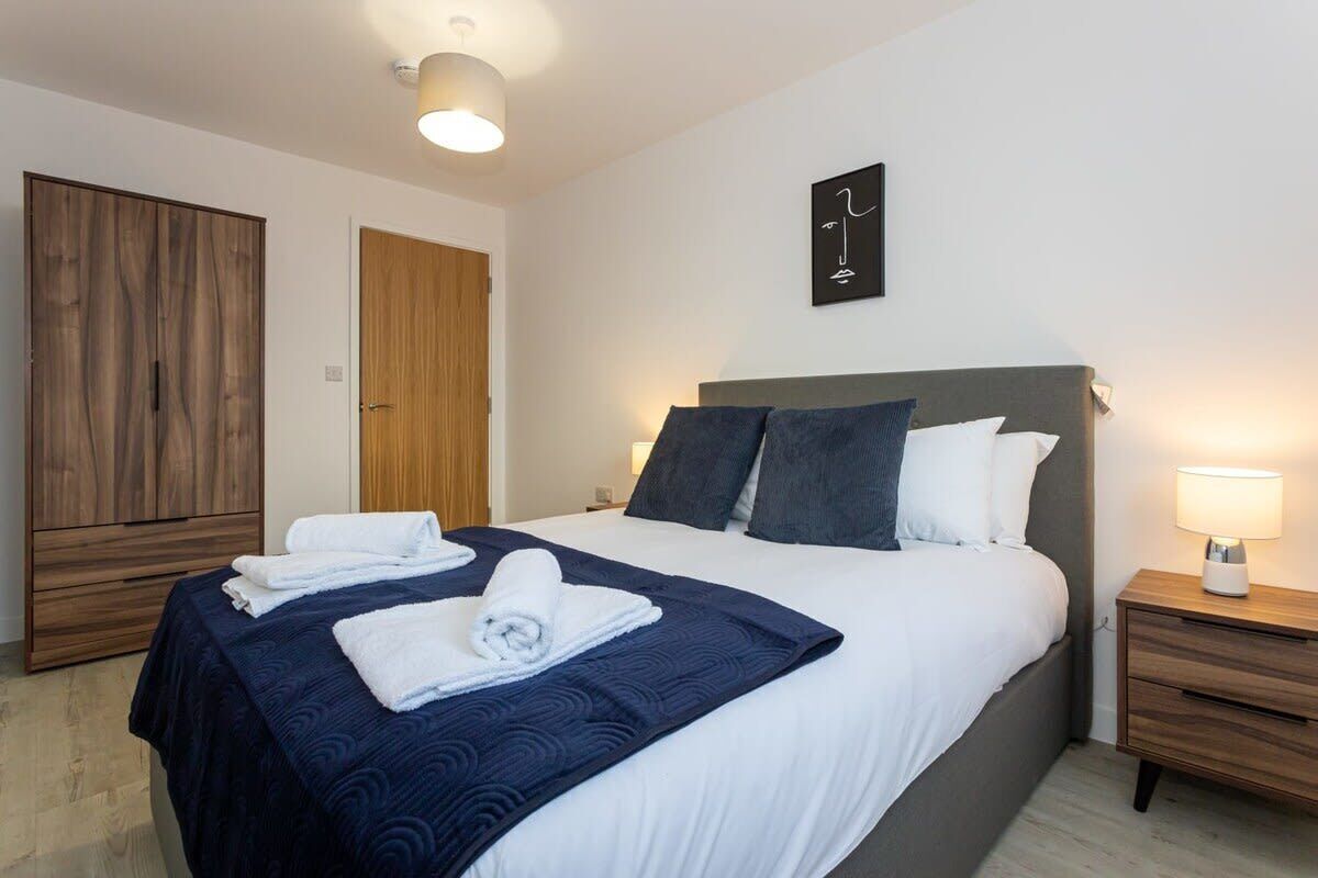 A neatly made bed with a dark blue blanket and white linens, a bedside lamp, and a wooden wardrobe.
