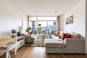 Een moderne woonkamer met een bank, een salontafel en een televisie. Er staan planten en er is uitzicht op de stad door grote ramen.