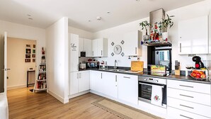 Una cucina moderna con mobili bianchi, un piano di lavoro nero e un forno integrato. Si nota una libreria con diversi oggetti e un orologio decorativo a parete.