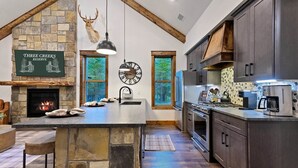 A modern kitchen with a stone island, wooden beams, a fireplace, and a view of the outdoors. - Pickleball, Pool, Splash Fountain ,Clubhouse , Walking Trails , Basketball (Broken Bow)