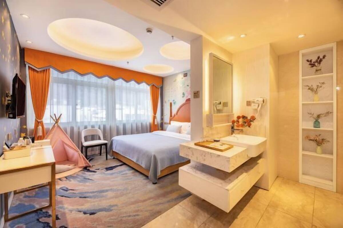 Ein Hotelzimmer mit einem Bett, einem Schreibtisch, einem Schminktisch mit Spiegel, einem Fenster mit Vorhängen und einem an der Wand befestigten Regal mit Dekoration.