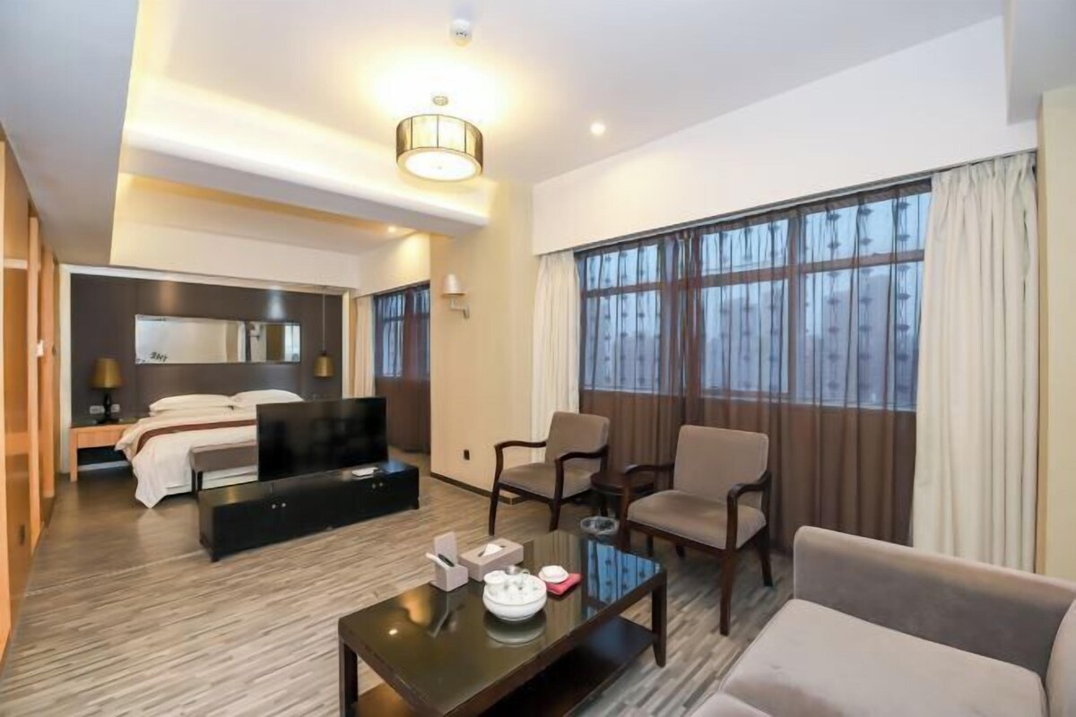 A hotel room with a bed, a TV, a seating area with chairs, a coffee table, and large windows with curtains.
