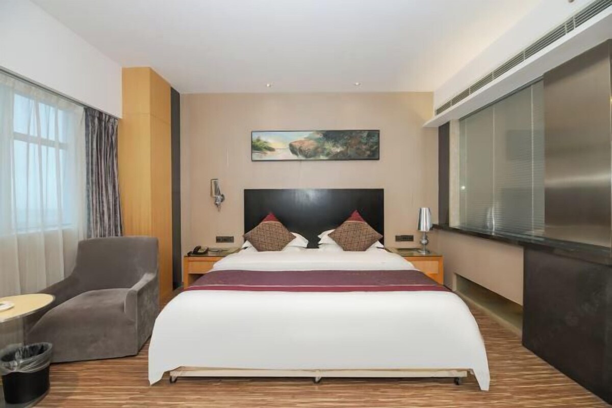 A hotel room with a large bed, a gray armchair, a small table, and a painting on the wall.