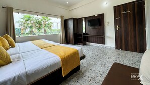 A hotel room with a bed, a TV, a wardrobe, and a balcony with a view of palm trees. - Hotel Vrinda Sarovar (Mathura)