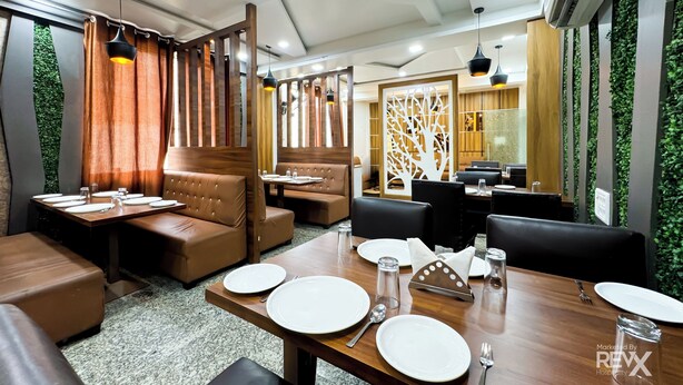A restaurant with wooden tables, leather booths, and a decorative tree panel. - Hotel Vrinda Sarovar (Mathura)