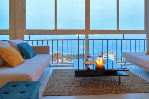 A modern living room with a large window, a sofa, a coffee table with a candle, and a view of the city.