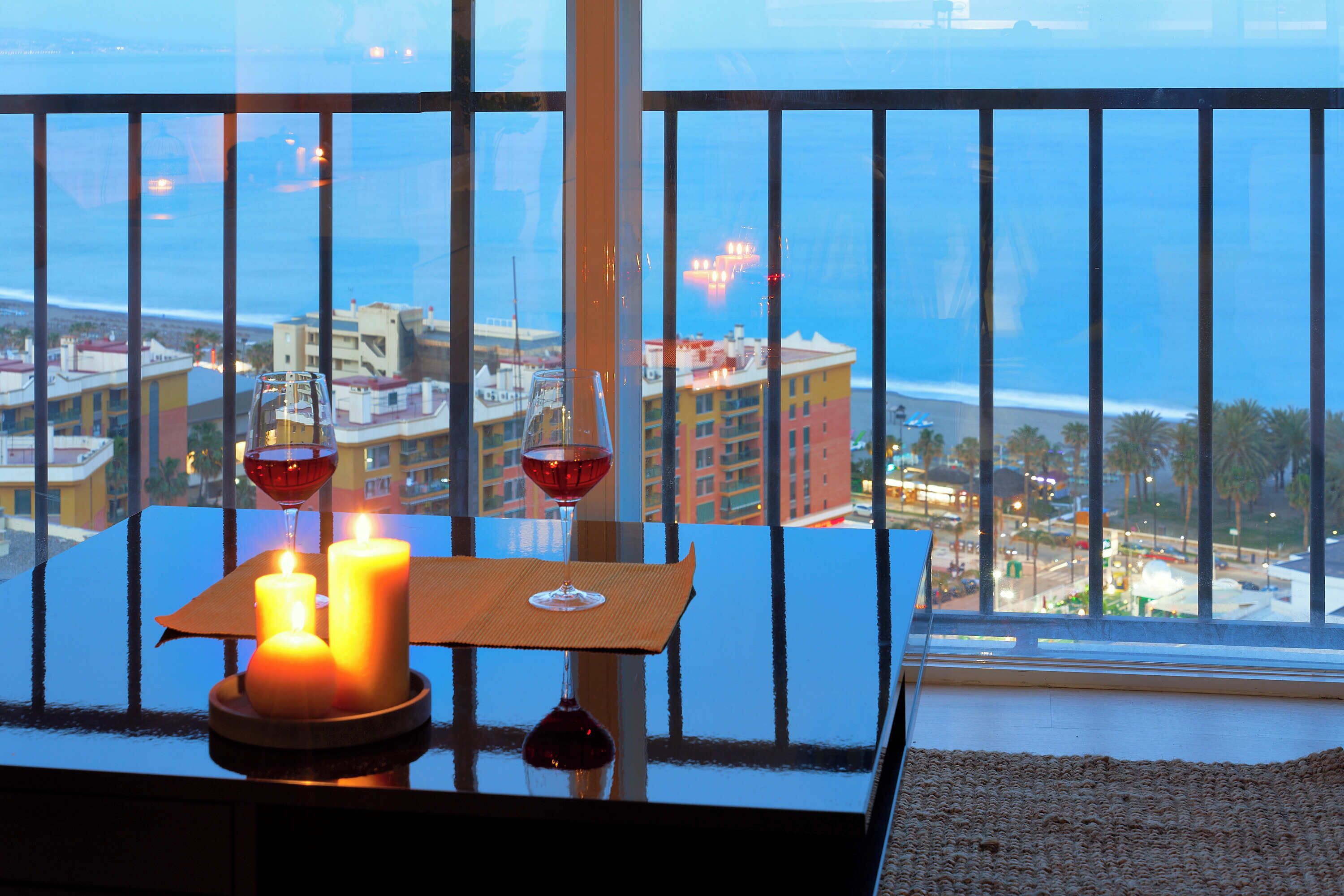 Une table éclairée à la bougie, avec un verre de vin rouge, offrant une vue sur le paysage urbain et l’océan.