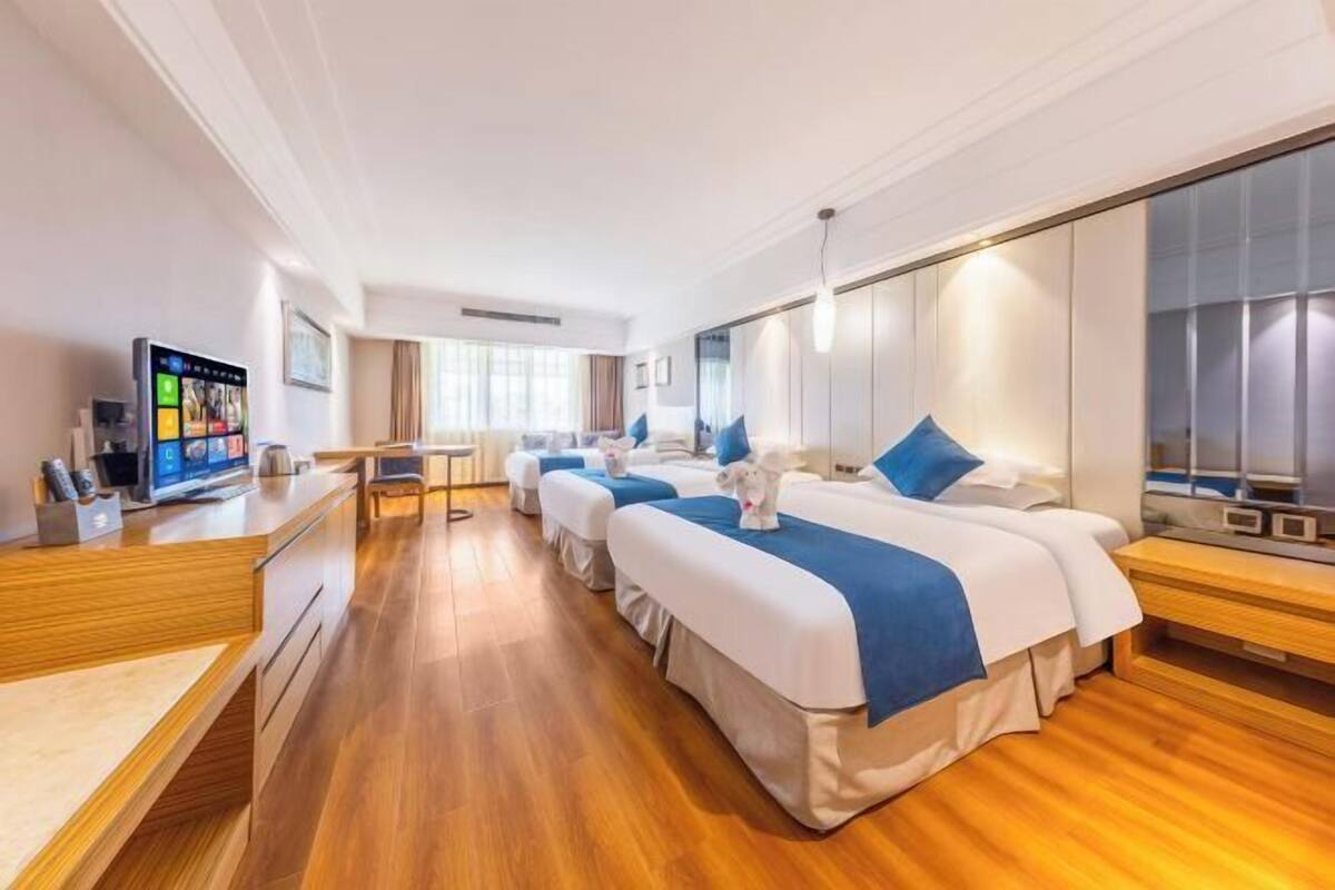 A hotel room with a wooden floor, three beds with blue and white bedding, a desk with a computer, and a large mirror.