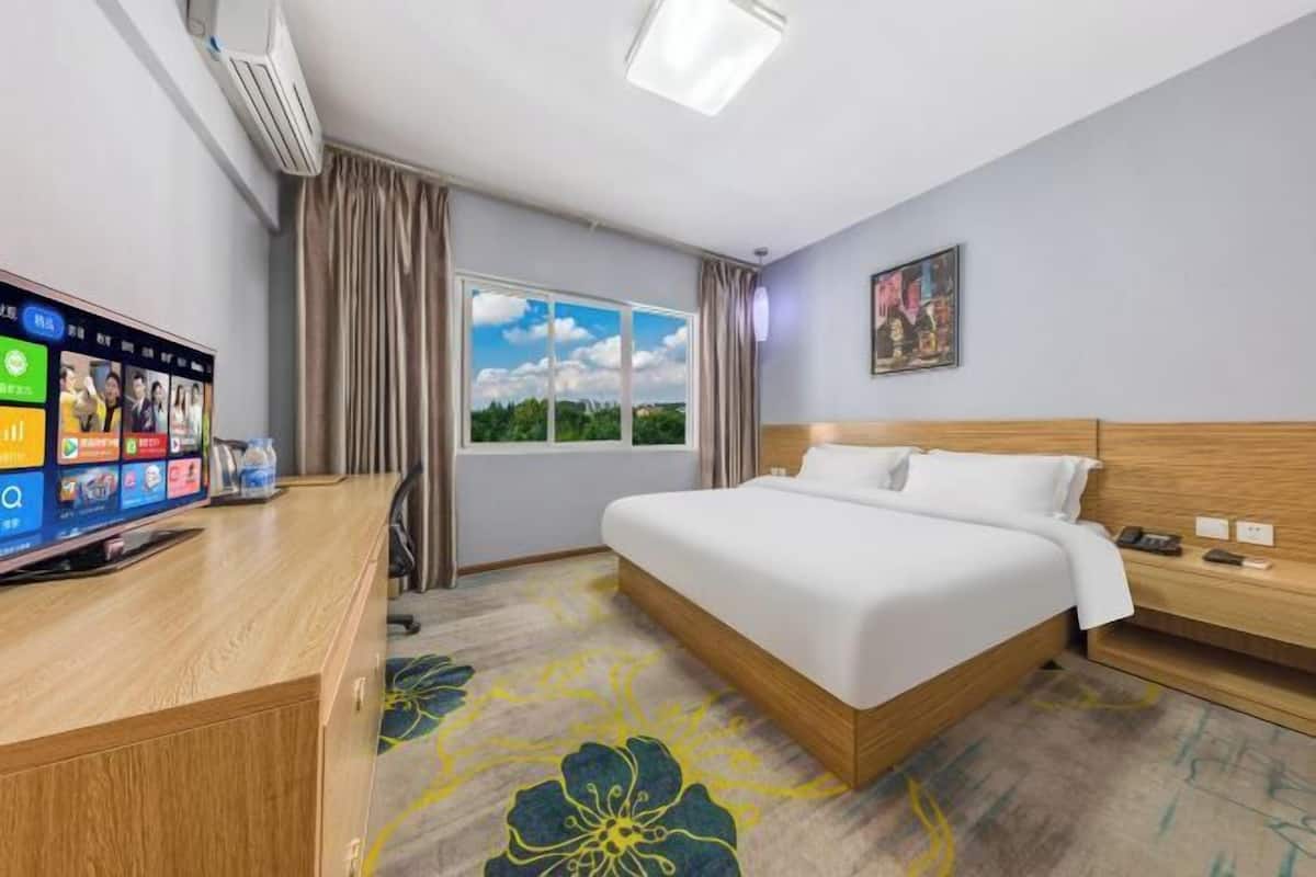 A hotel room with a bed, a desk with a computer, a window with curtains, and a painting on the wall.
