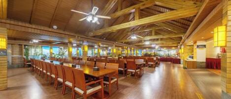 A spacious indoor dining area with high ceilings and wooden beams, featuring multiple tables and chairs arranged for groups.