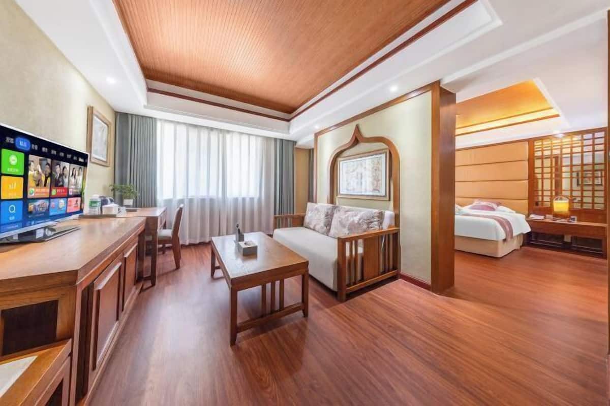 A hotel room with a wooden floor, a sofa, a bed, a desk with a TV, and a window with curtains.