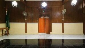 Una habitación con piso de baldosas negras, paredes de madera y un acceso central con cortina naranja. Se observan dos ventiladores de pared y dos lámparas colgantes.