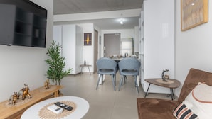 Een moderne woonkamer met een flatscreen televisie, een eethoek met twee stoelen, een klein tafeltje met een decoratief object, en een bruine bank met een gestreept kussen.