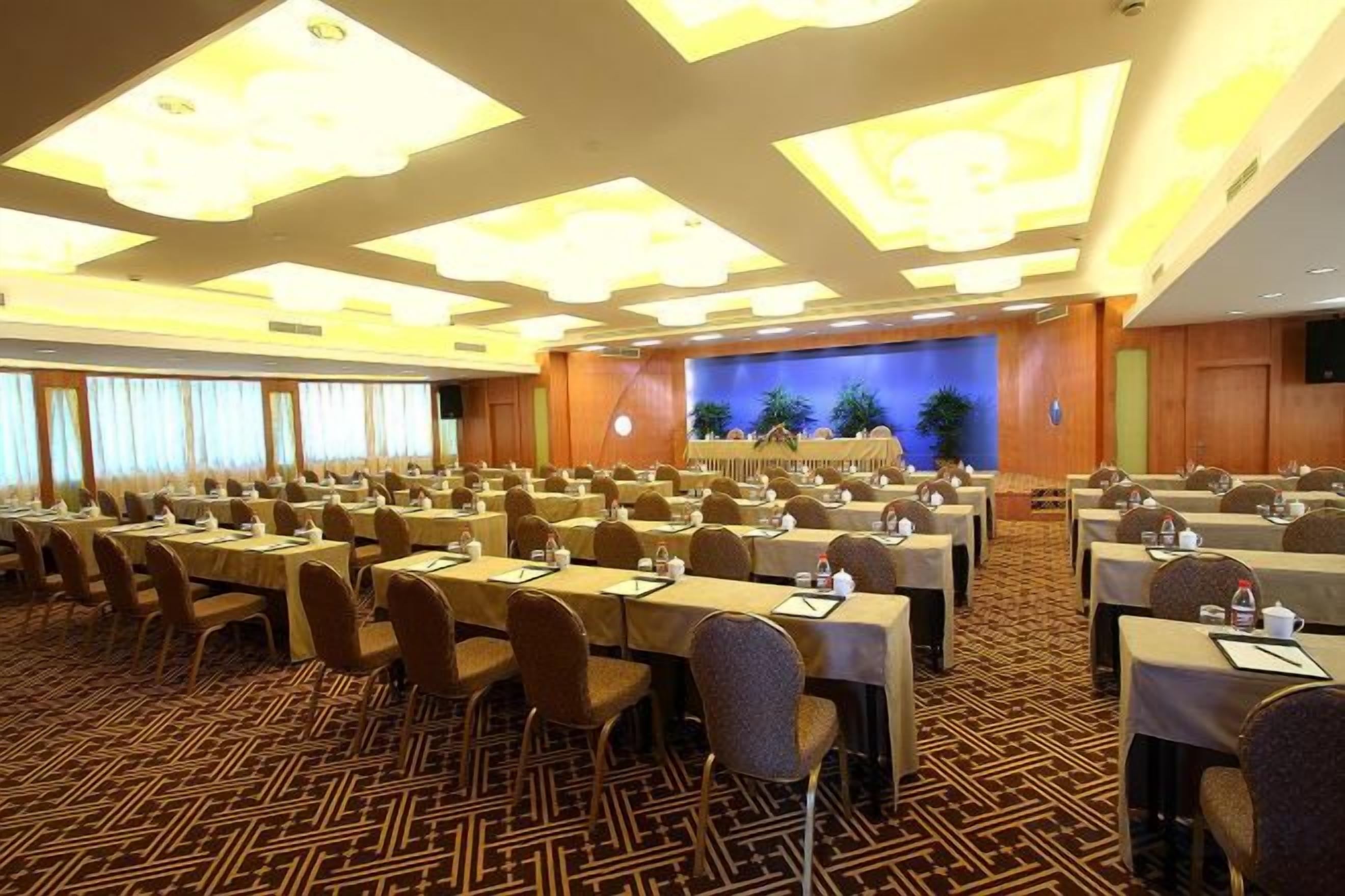 A conference room with multiple tables and chairs, a stage with a blue backdrop, and a patterned carpet.