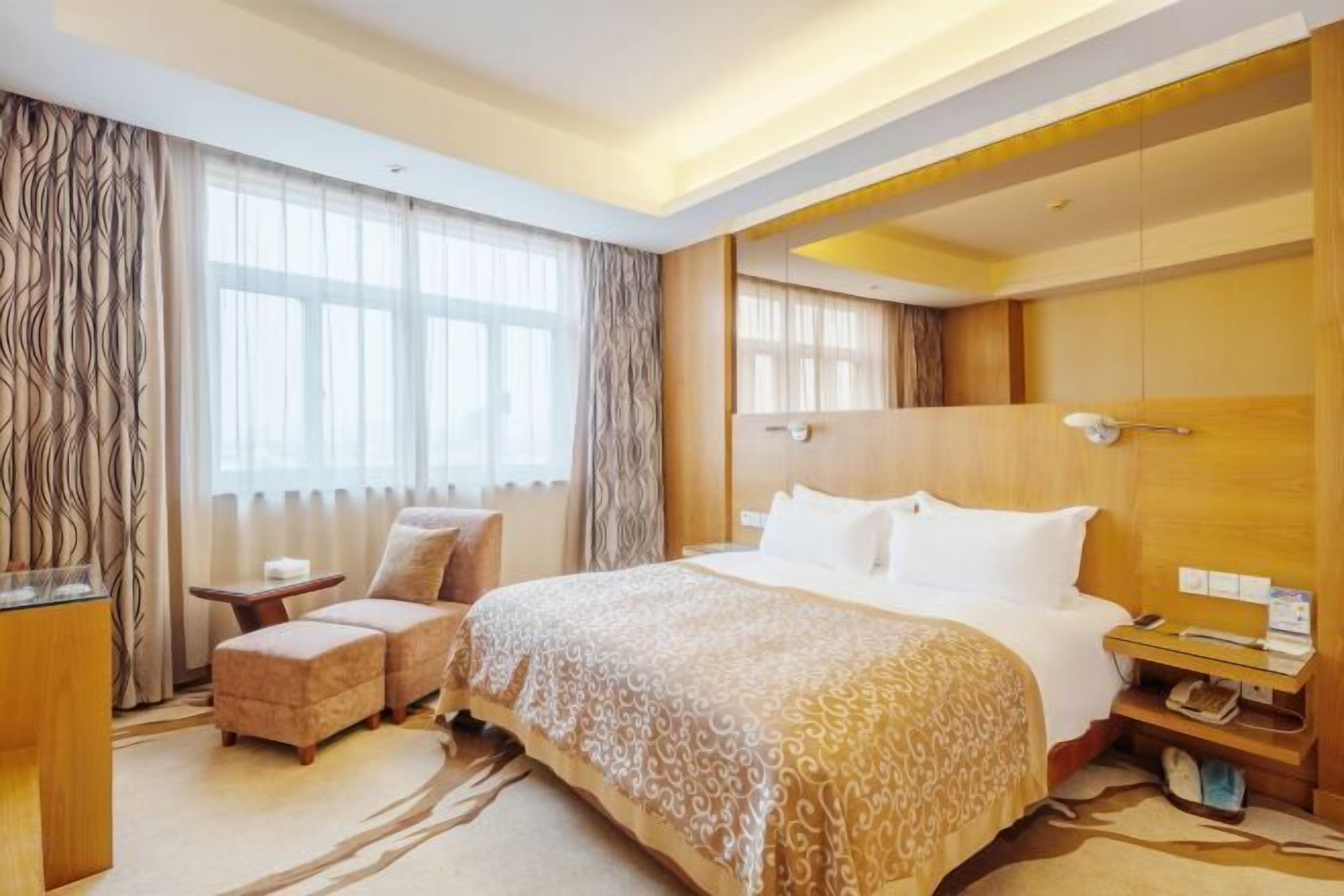 A hotel room with a large bed, a sitting area with a chair and ottoman, a bedside table, and a window with curtains.