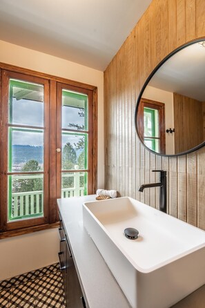A bathroom with a white sink, wooden panel walls, a large mirror, and a window with a view of mountains. - Swiss Chalet with Private Deck & Mountain Views (Golden)