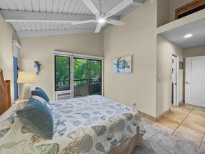 A bedroom with a bed, a ceiling fan, a window with a view of greenery, a door, and a wall with a turtle painting.