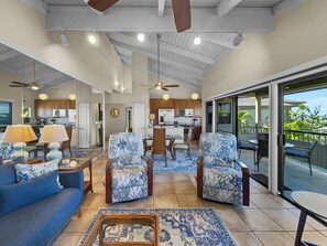 A spacious living area with a large sofa, two patterned armchairs, a coffee table, and a dining table with chairs. There is a kitchen area with wooden cabinets and a ceiling fan. The space opens to a balcony with outdoor seating.