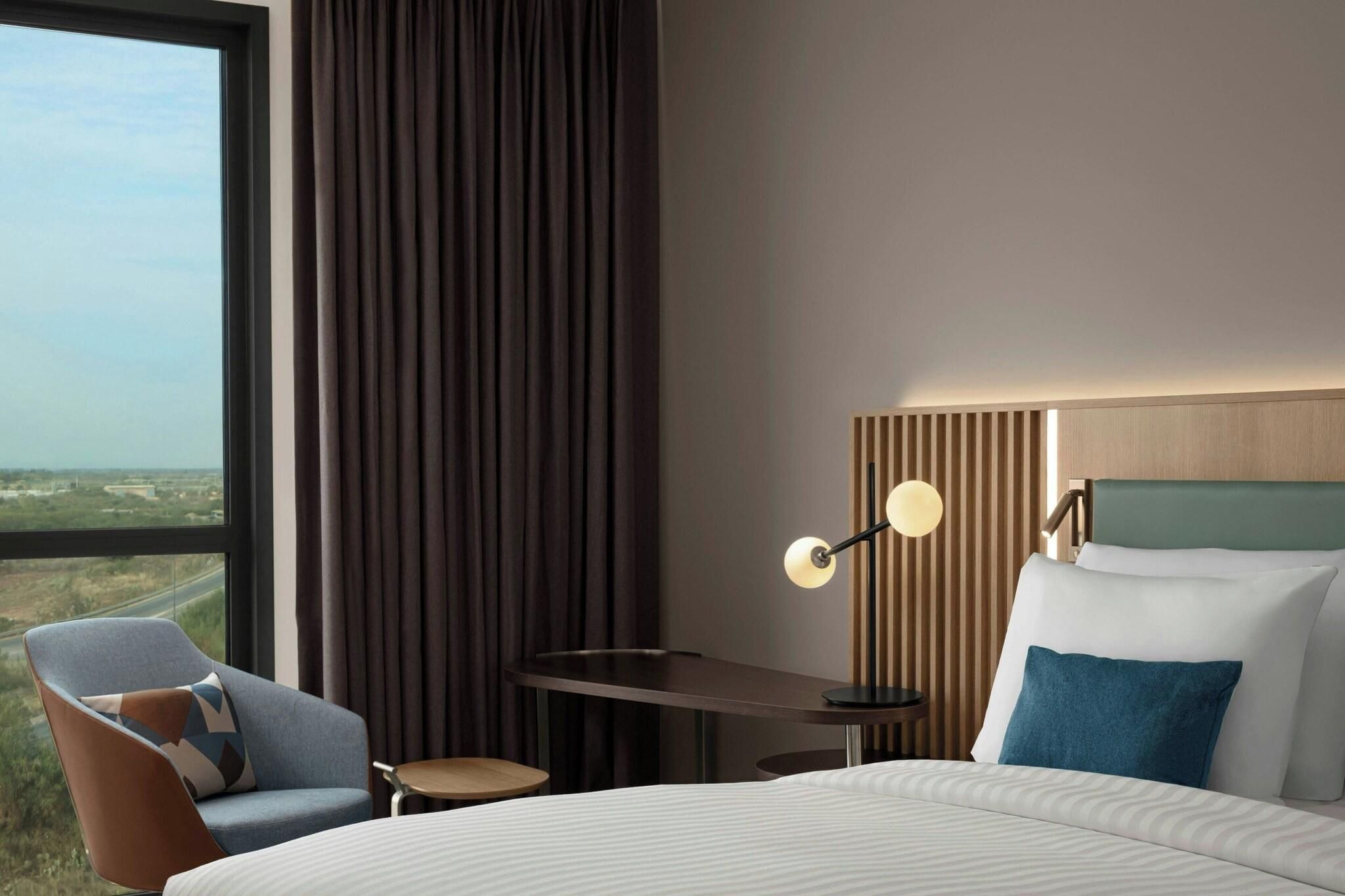 A hotel room with a large window, a bed, a desk, a chair, and a lamp.