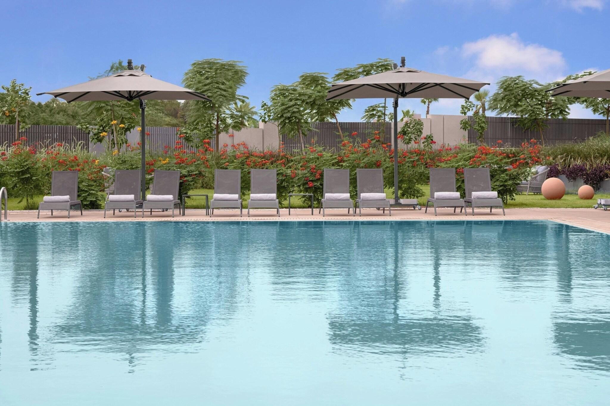 Une piscine entourée de verdure et d’une clôture, avec des chaises longues et des parasols.