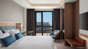 Een moderne hotelkamer met een groot bed, een zithoek met een stoel en bankje, een kleine tafel en een groot raam met uitzicht op de stad.
