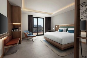 A modern hotel room with a large bed, a seating area with a chair, a TV, and a view of the city through the window. - Courtyard By Marriott Dakar Diamniadio (Diamniadio)
