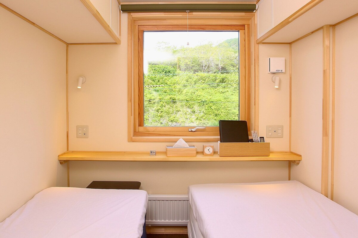 Ein kleines Hotelzimmer mit einem Bett, einem Fenster mit Blick auf Grünflächen, einem Nachttisch mit einer Uhr und einer Steckdose.