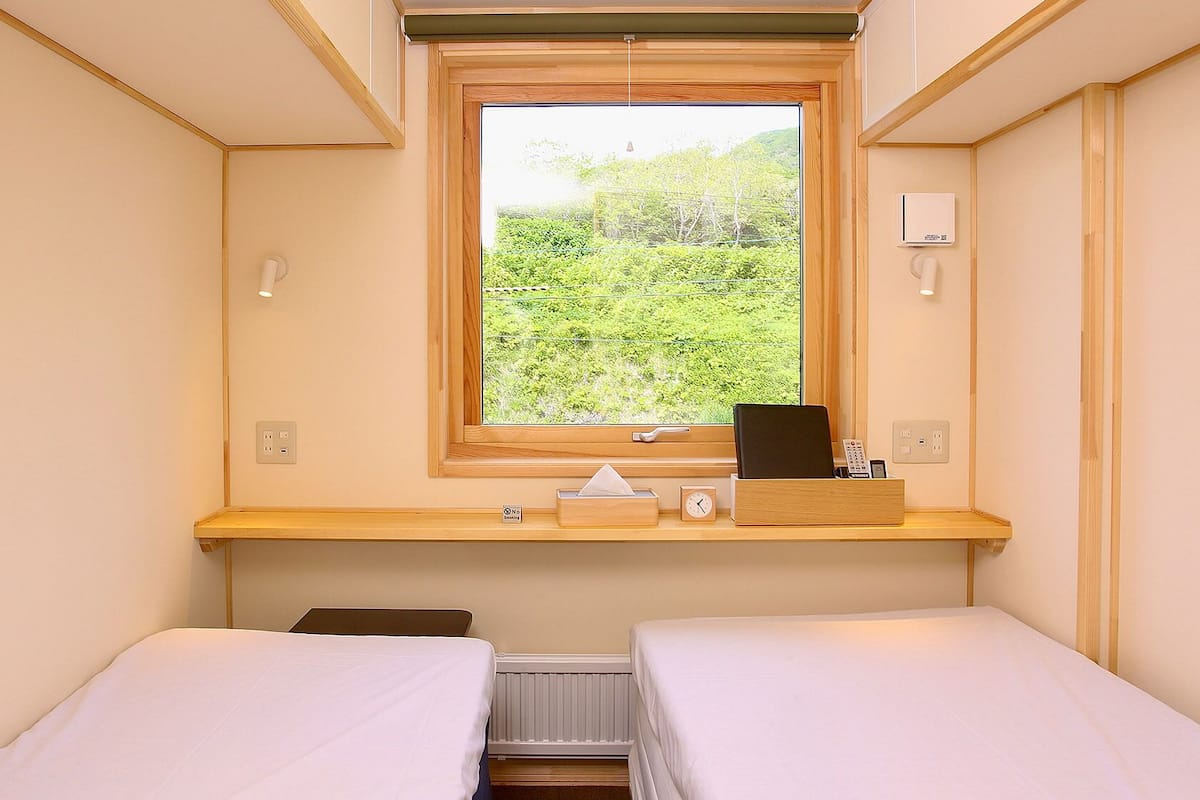 A small, well-lit room with two beds, a window with a view of greenery, a bedside table with a clock, and a power outlet.