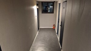 A narrow, dimly lit hallway with a grey carpet, a window with curtains, and a fire extinguisher on the wall.