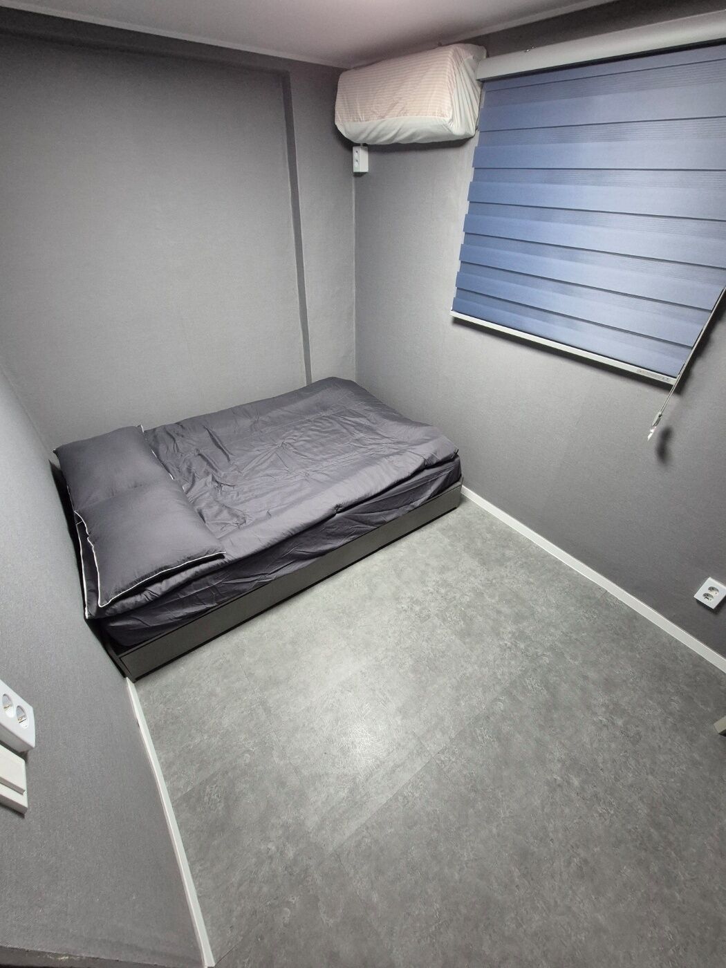 Quarto pequeno e compacto com uma cama de solteiro, janela com persianas e ar condicionado fixado na parede.