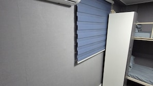 A compact room with a built-in air conditioner, a window with blinds, and a bunk bed.