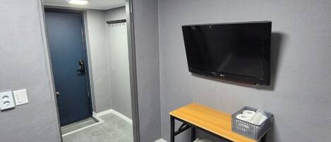 A room with a grey carpet, a wooden desk with a stool, a television mounted on the wall, and a door leading to another room.