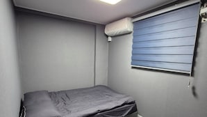 A small, single-bed room with a grey mattress, a ceiling light, a window with blinds, and a wall-mounted air conditioner.