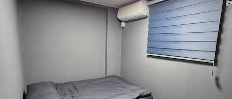 A small, single-bed room with a grey mattress, a ceiling light, a window with blinds, and a wall-mounted air conditioner.