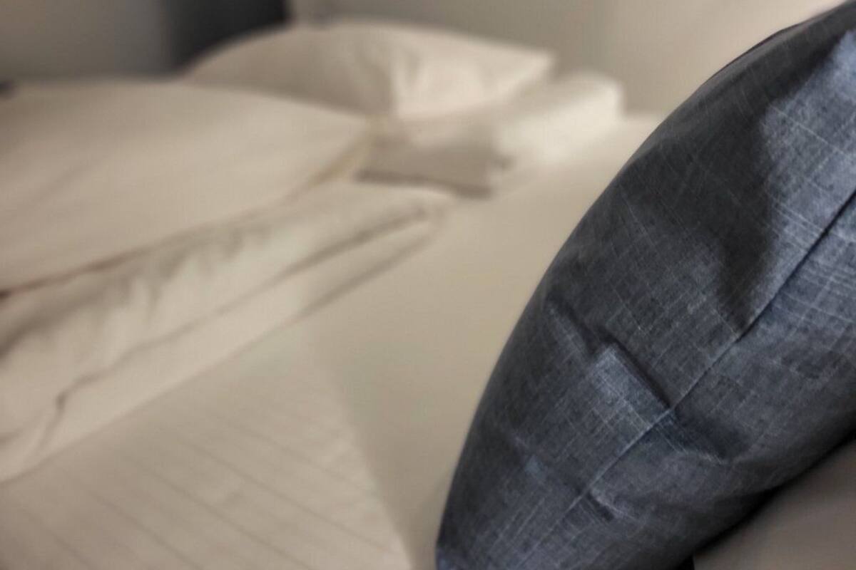 Een bed met een wit laken en een grijs kussen.