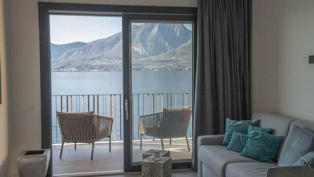 A living room with a sofa and two chairs, a balcony with a view of a lake and mountains, and a sliding glass door. - Attilio High Living Suites - Adults Only (Torri del Benaco)
