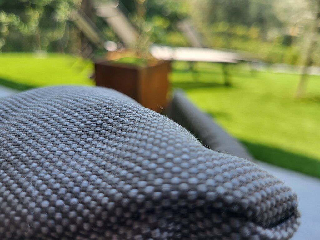 A close-up of a textured fabric, possibly a blanket or cushion, with a blurred outdoor background suggesting a garden or park setting.