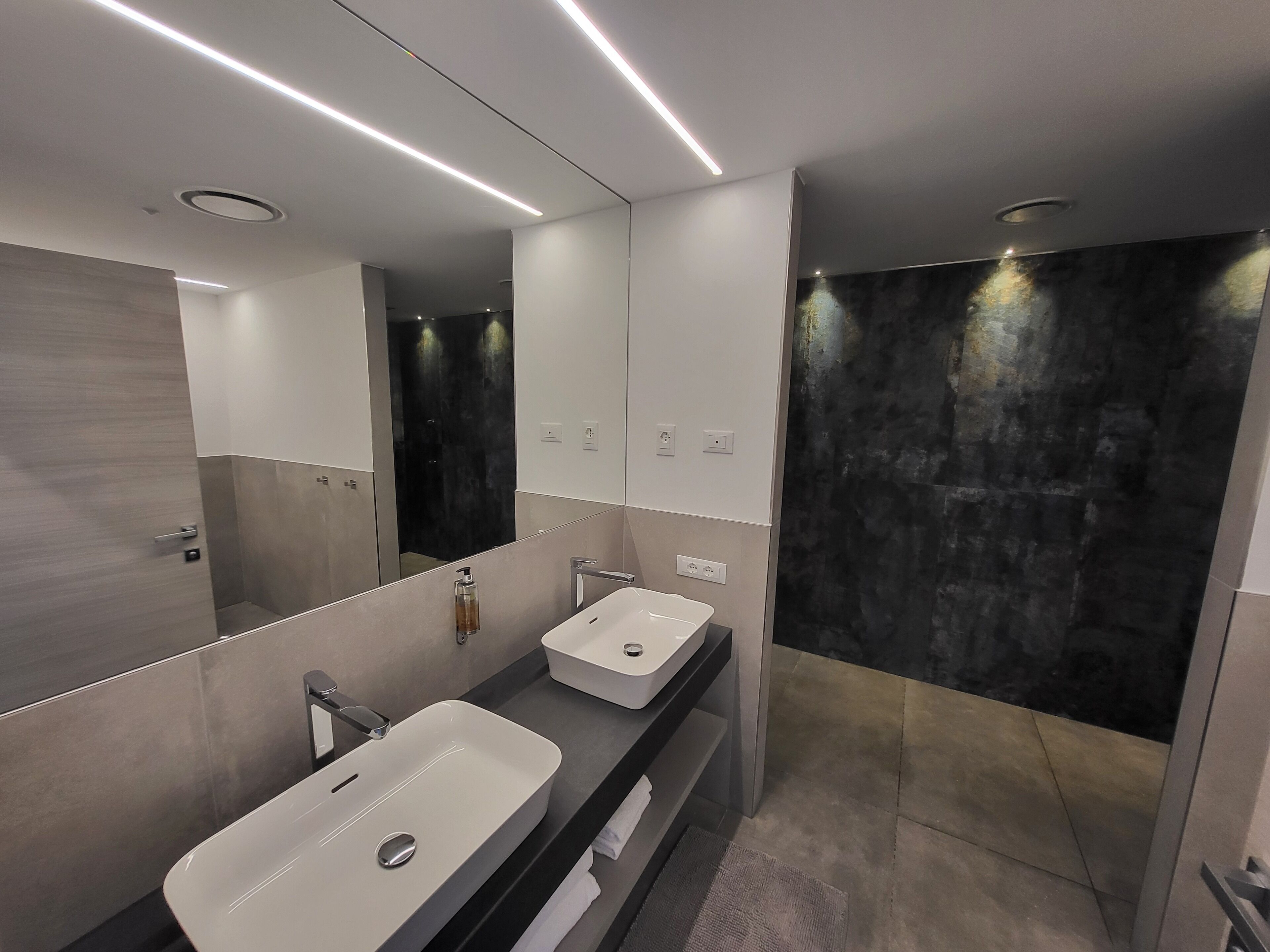 A modern bathroom with two sinks, a large mirror, and a dark shower area.