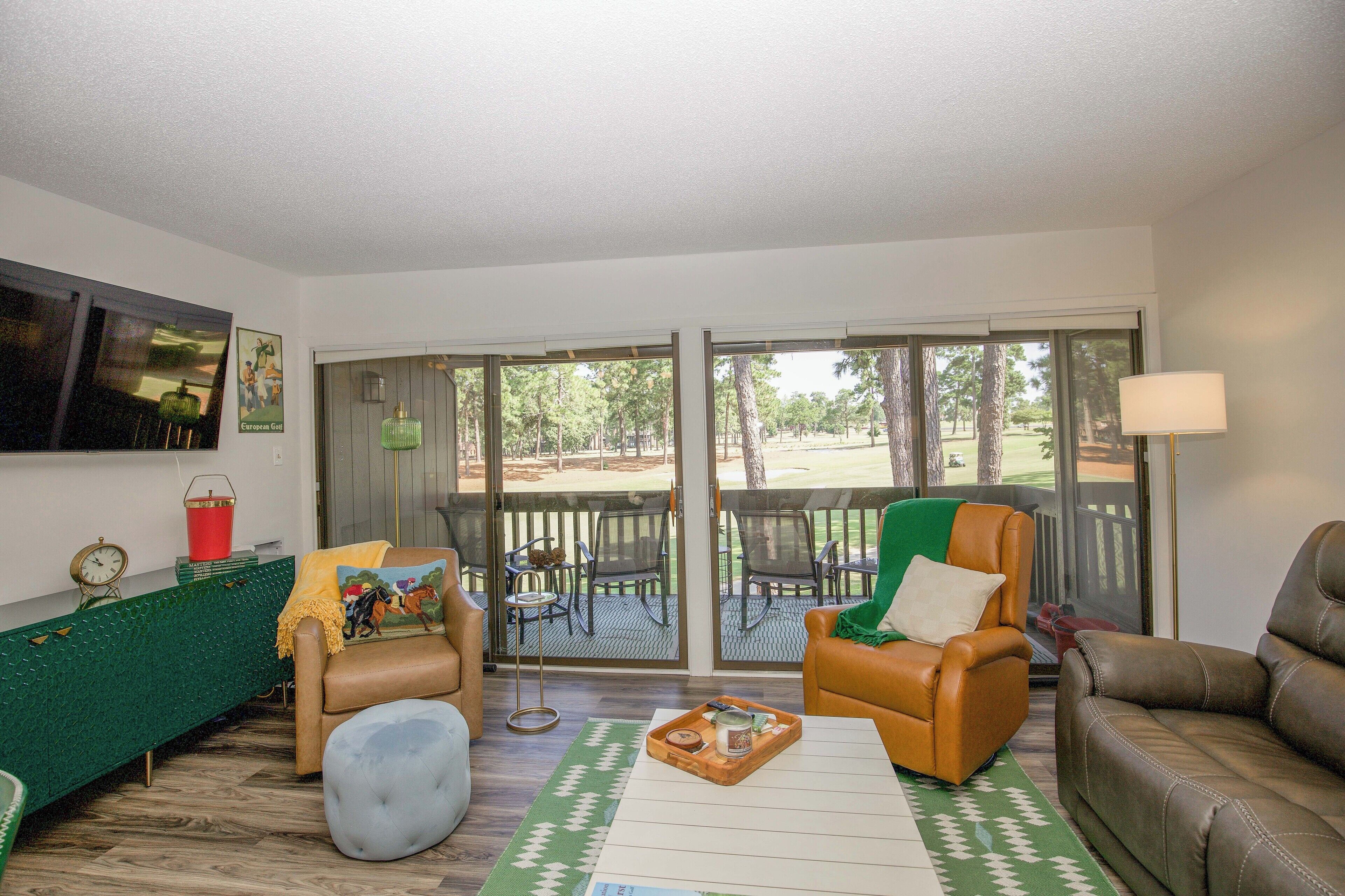 A living room with a green sofa, a brown leather couch, and a coffee table. There is a sliding glass door leading to a patio with outdoor furniture.