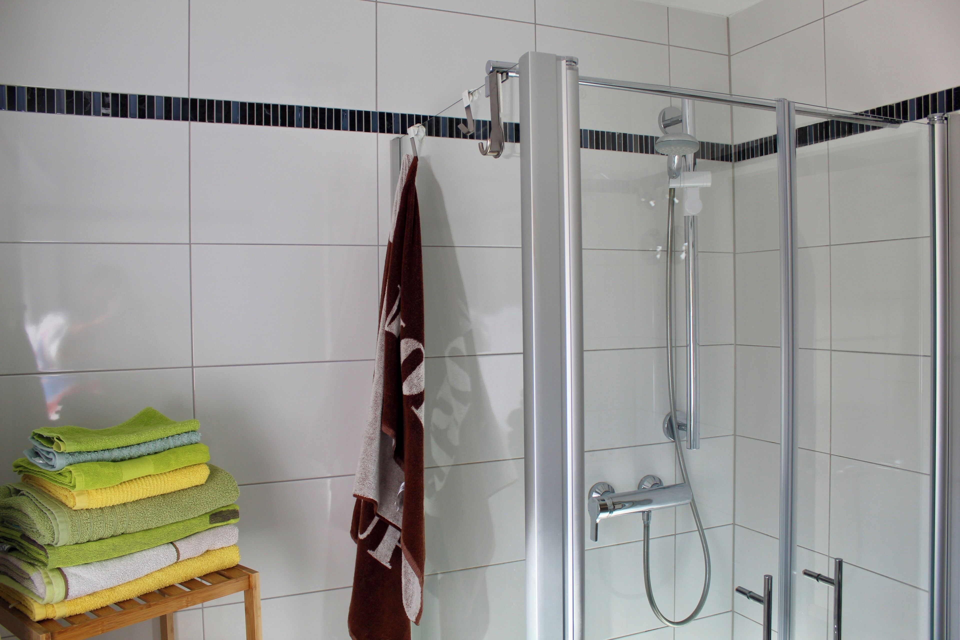Una ducha envuelta en vidrio con un cabezal de ducha de mano, una pila de toallas dobladas sobre un taburete y un perchero contra una pared de azulejos.