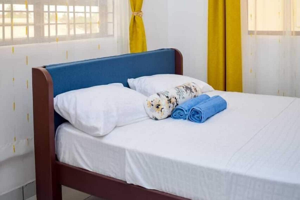 Ein Einzelbett mit weißer Bettwäsche, blauen Kissen, gelben Vorhängen und einem Holz-Kopfstück.