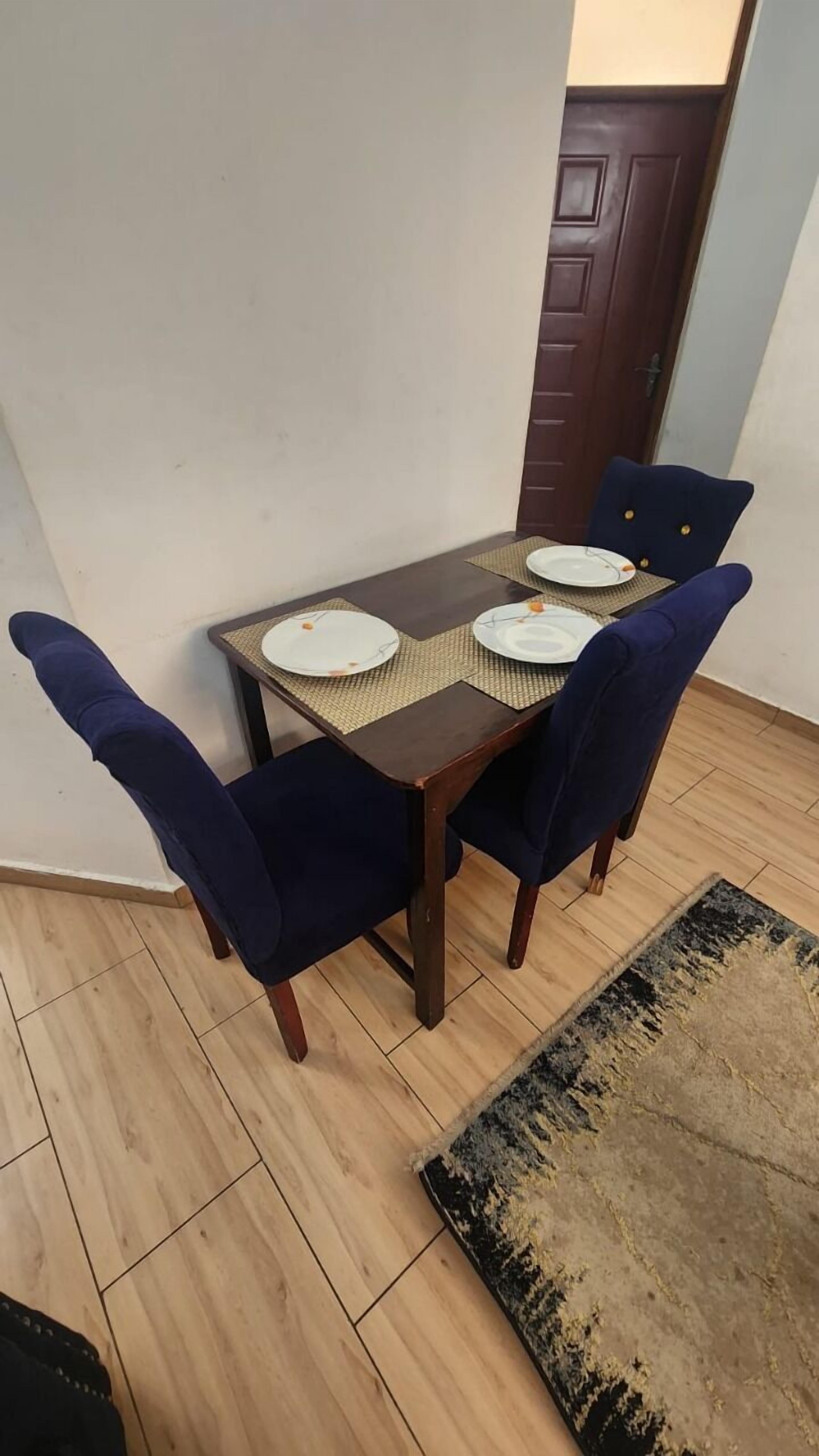 Een eettafel met vier stoelen, een geweven placemat en een donker houten deur op de achtergrond.