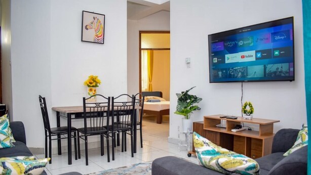 A modern living room with a TV, a dining area with a table and chairs, and a sofa. - Casey's Apartments Bamburi (Mombasa)