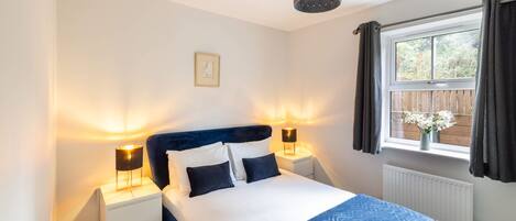 A bedroom with a bed, two bedside lamps, a window with curtains, a radiator, and a hanging light fixture.