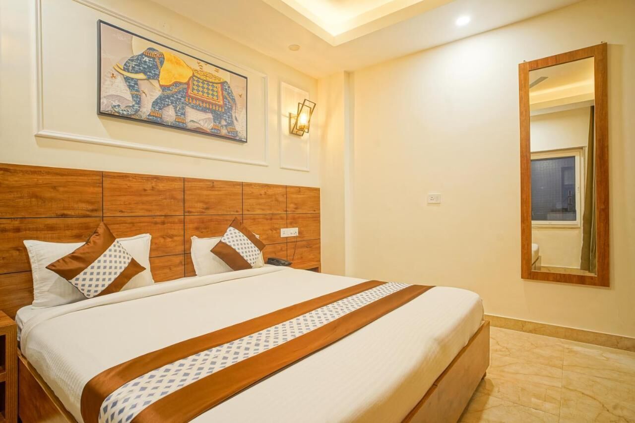 A hotel room with a wooden headboard, a large bed with patterned bedding, a wall-mounted artwork, a wall-mounted light fixture, and a mirror.
