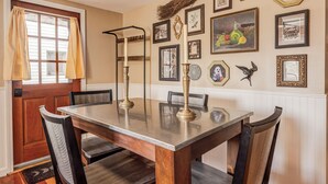 A dining room with a wooden table and chairs, a window with curtains, and various framed pictures on the wall.