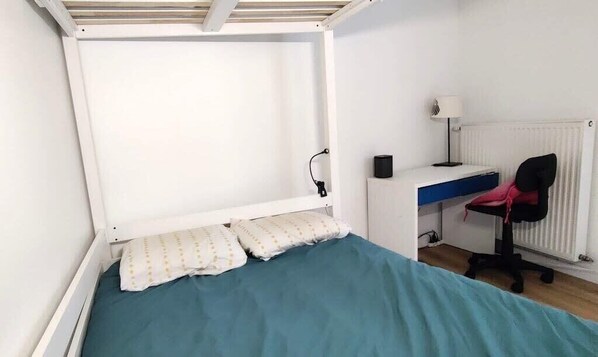 Ein kleines Zimmer mit einem Einzelbett, einem Schreibtisch, einem Stuhl, einem Heizkörper und einer Lampe.