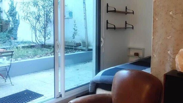 Un salon doté d’une porte-fenêtre coulissante donnant sur un balcon, d’un fauteuil en cuir marron avec un coussin bleu et d’une petite table accompagnée d’une chaise.