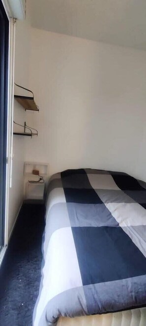 Ein kleines, ordentlich bezogenes Bett mit karierter Tagesdecke in einem Zimmer mit weißen Wänden und einem Fenster.